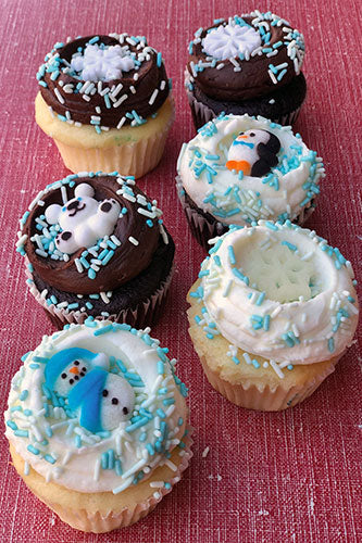 Six festive cupcakes decorated with white and chocolate frosting, blue and white sprinkles, and winter-themed toppers like snowmen and snowflakes.