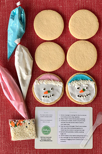 A cookie decorating kit with six sugar cookies, a pouch of royal icing, a garnish pouch, and a mini spatula, displayed on a red surface with a recipe card.