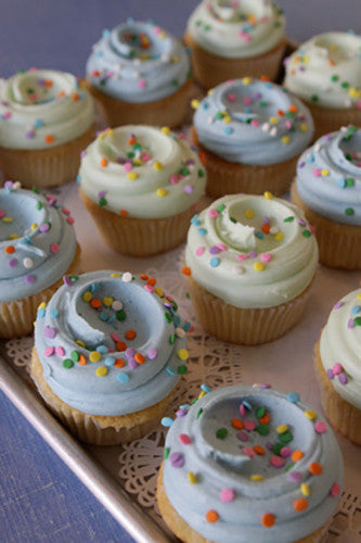 A tray of vanilla cupcakes with light blue and white buttercream frosting, topped with multicolored sprinkles.