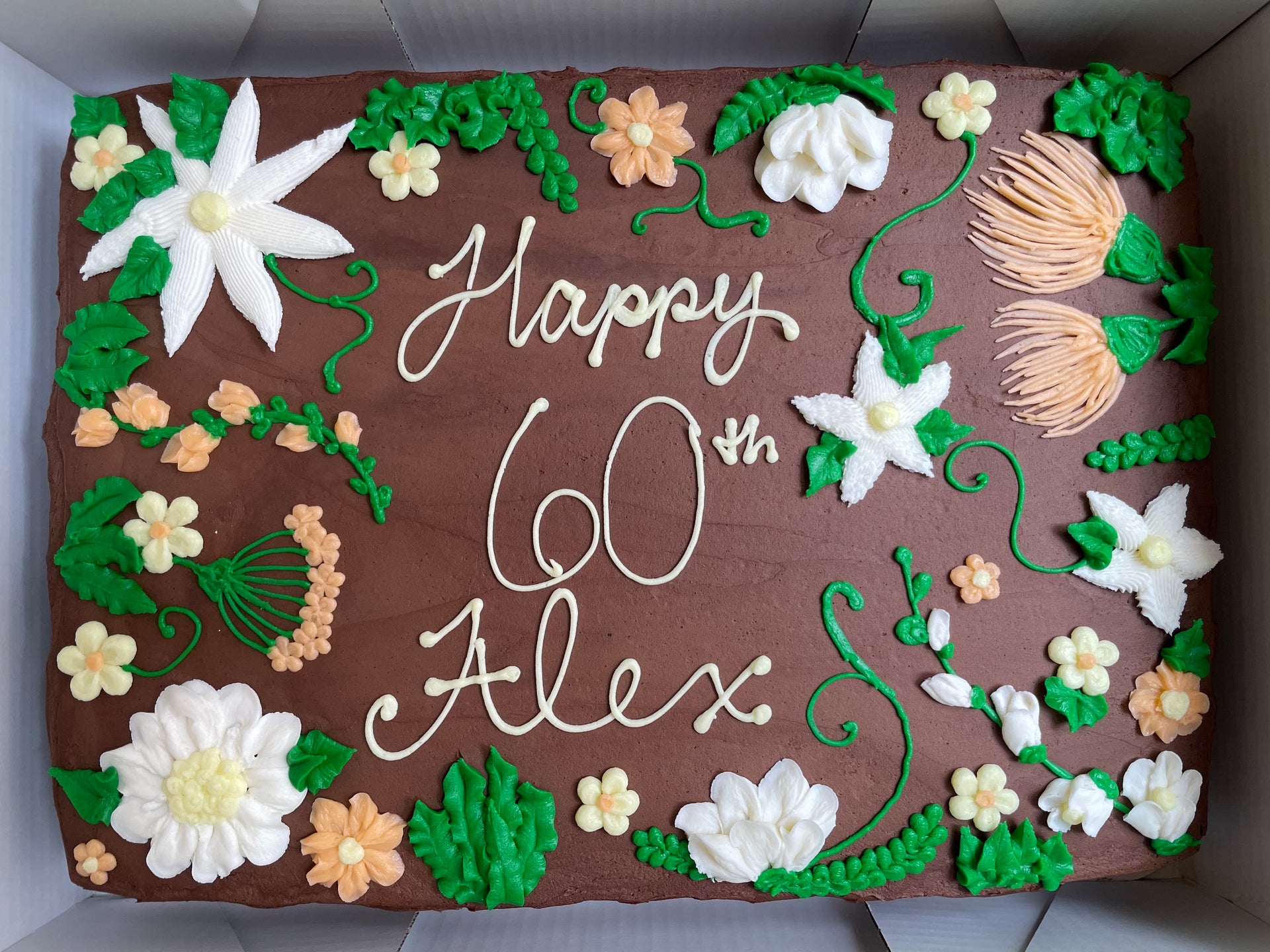 A rectangular chocolate cake decorated with white and yellow icing flowers, green leaves, and the words 'Happy 60th Alex' in white script, photographed from above inside a white box.