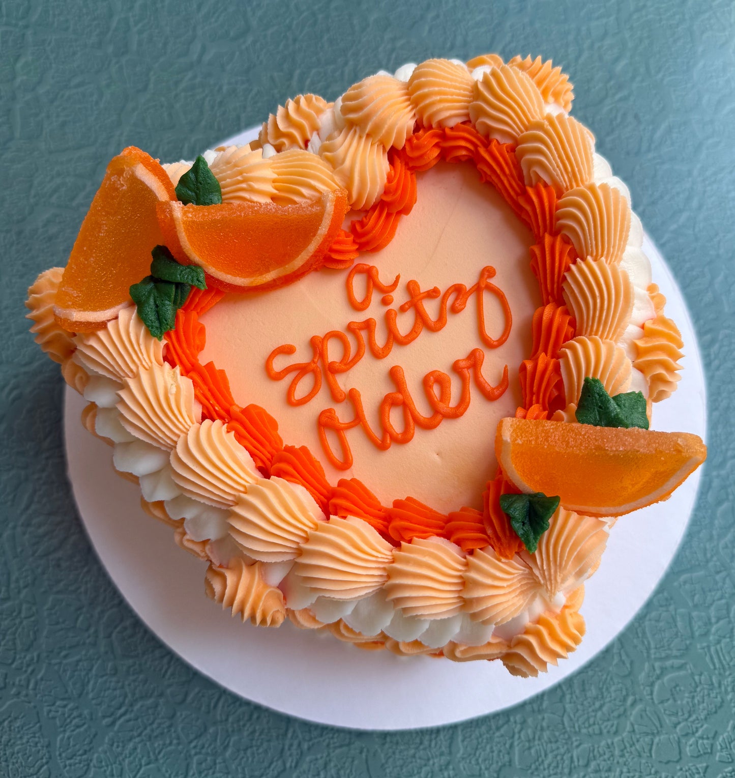 A decorated cake with orange buttercream frosting and candied orange wedges, with the text 'A Spritz Older' written on top.
