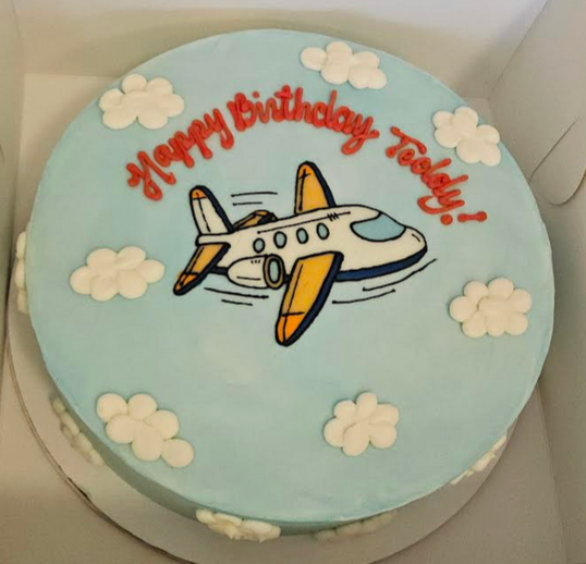 Birthday cake with airplane design and 'Happy Birthday Teddy!' text on a light blue background.