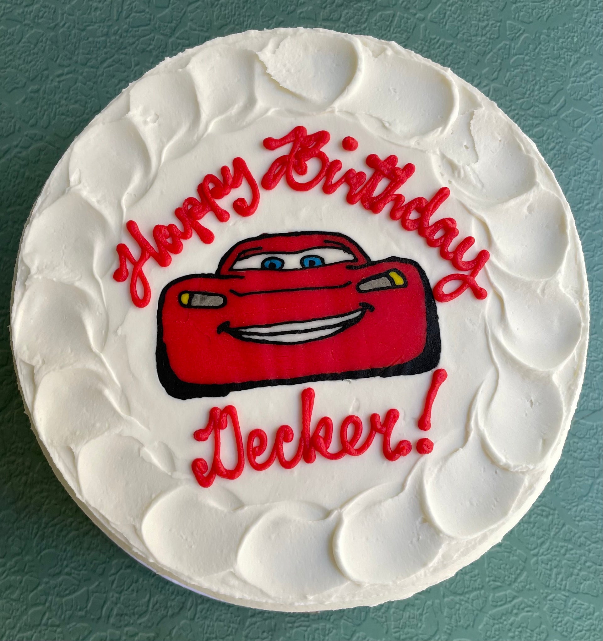 Round white cake with red car and 'Happy Birthday Decker!' text on a teal background