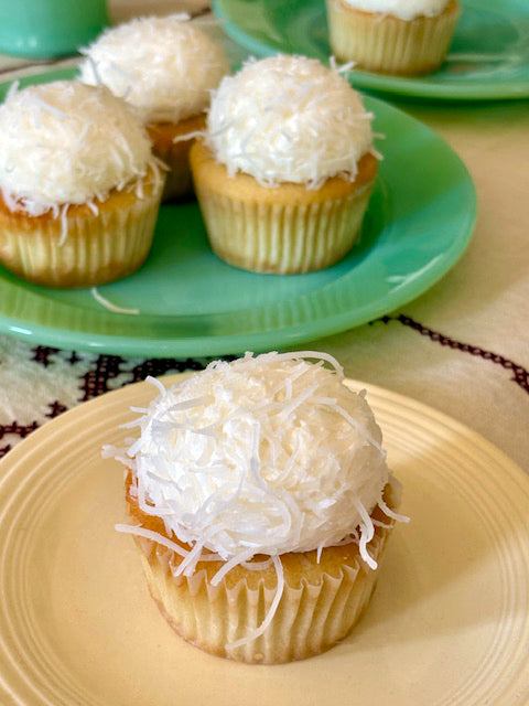 Cupcakes with coconut topping on a yellow plate, with more cupcakes in the background.
