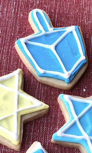 Sugar cookies decorated with Hanukkah themes, including a blue dreidel and a yellow Star of David, outlined in white icing, on a red background.