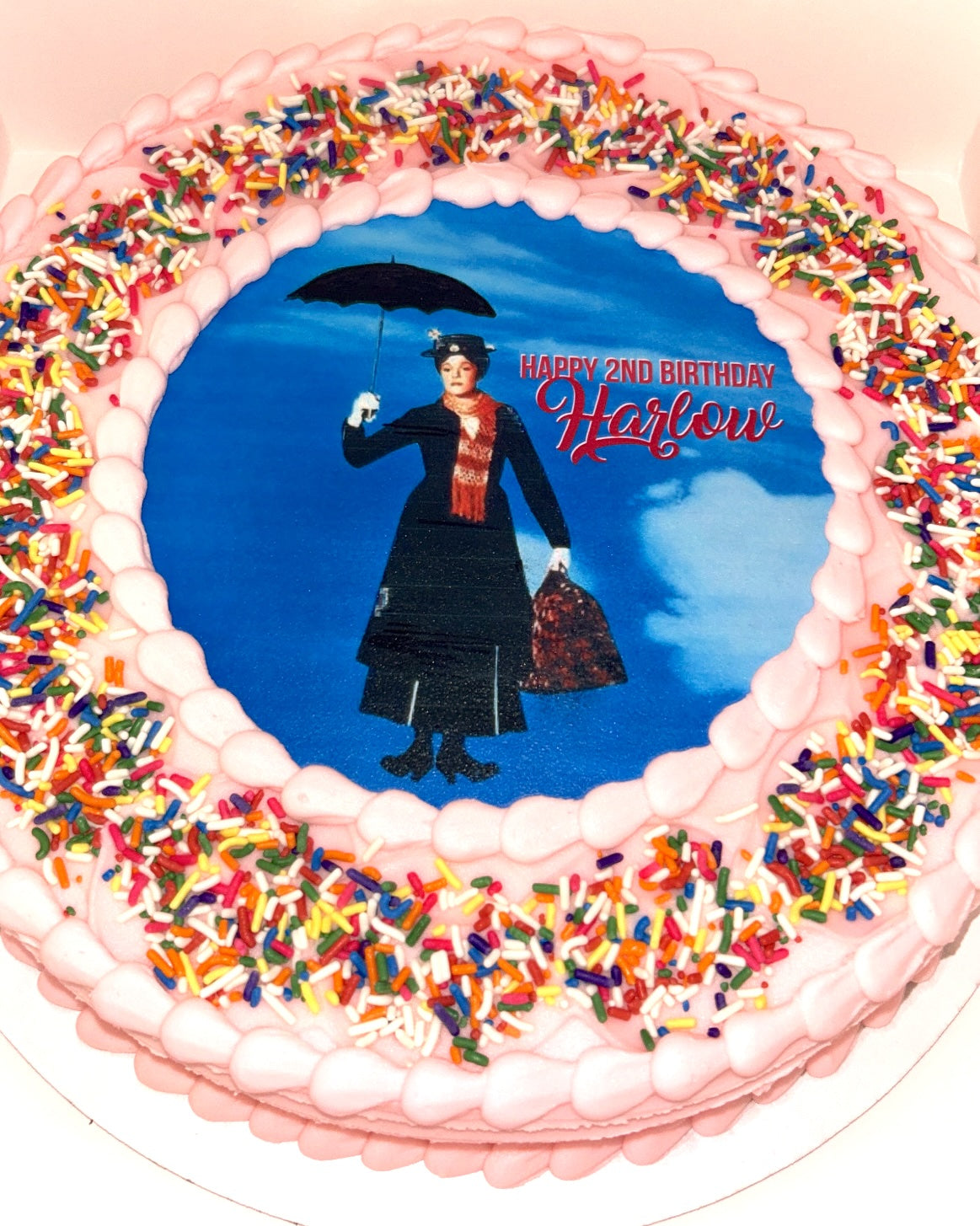 A vibrant, round cake frosted smoothly in a soft pink buttercream. The top of the cake features a large, circular edible image against a bright blue background, depicting the iconic character Mary Poppins, dressed in her classic black coat, red scarf, and hat, holding her open black umbrella and carpet bag. To the right of Mary Poppins, in white and red script, are the words "HAPPY 2ND BIRTHDAY Harlow" and there is a rainbow sprinkles border.