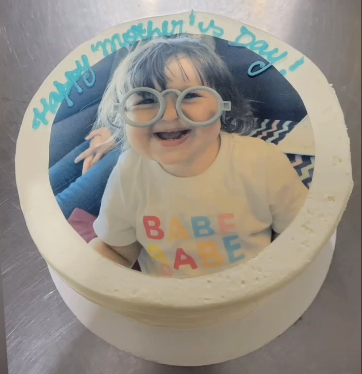 A round, white frosted cake with a top surface adorned with a large, circular edible image of an adorable, smiling toddler. The child has dark hair and is wearing a white t-shirt with colorful letters spelling out "BABE". They are playfully wearing light-colored, possibly blue or white, novelty glasses with large round frames. Above the edible image it says 'Happy Mother's day!'