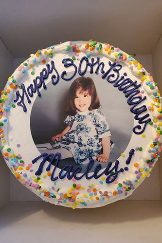 This cheerful image displays a round cake, generously frosted in smooth white buttercream. The top of the cake features a large, circular edible image of a young girl with brown hair, smiling sweetly while wearing a patterned light-colored dress with blue floral designs. She appears to be sitting or posing on the floor.