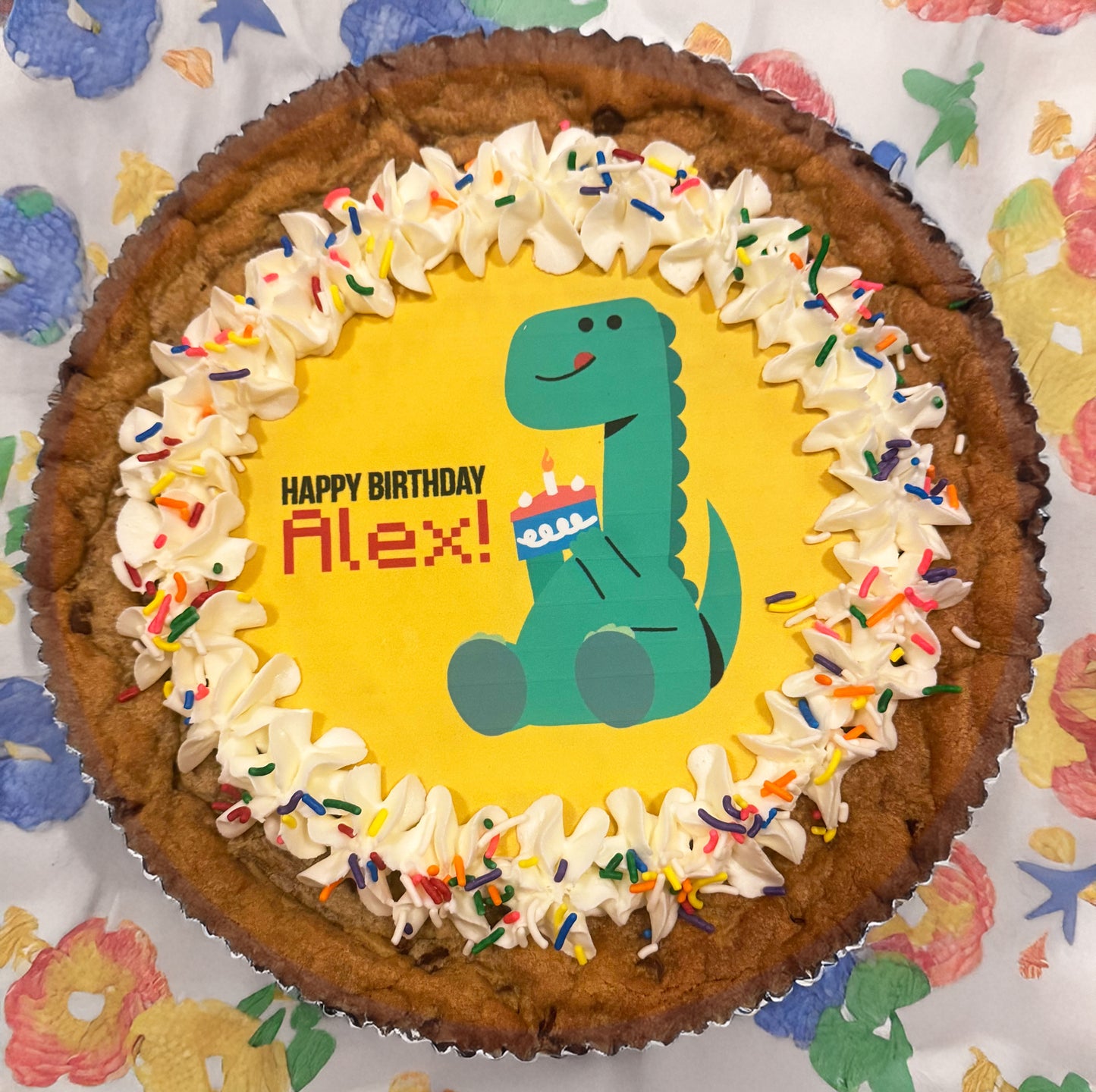 Cookie cake with dinosaur design and 'Happy Birthday Alex!' text on a colorful patterned background