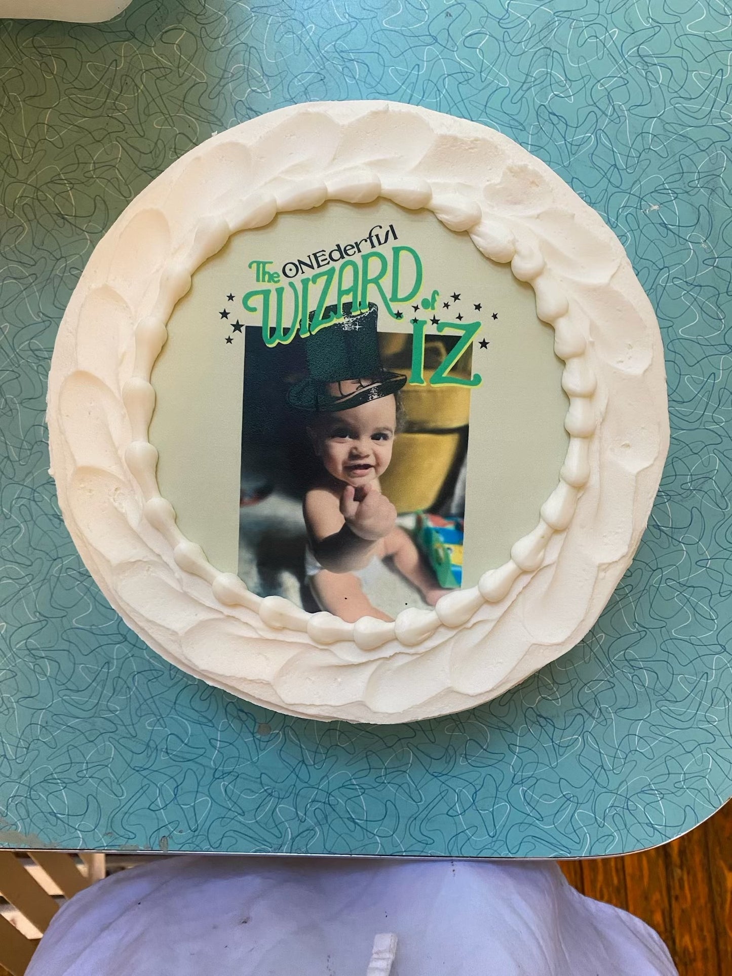 A round cake, generously covered in a thick, textured white frosting, giving it a soft, cloud-like appearance. The top of the cake showcases a circular edible image with a light green background. At the center of this edible image is a charming photograph of a baby, seemingly in a diaper, joyfully looking towards the camera and holding up one finger. The baby is wearing a whimsical green top hat, adorned with a buckle, reminiscent of a leprechaun or wizard's hat.