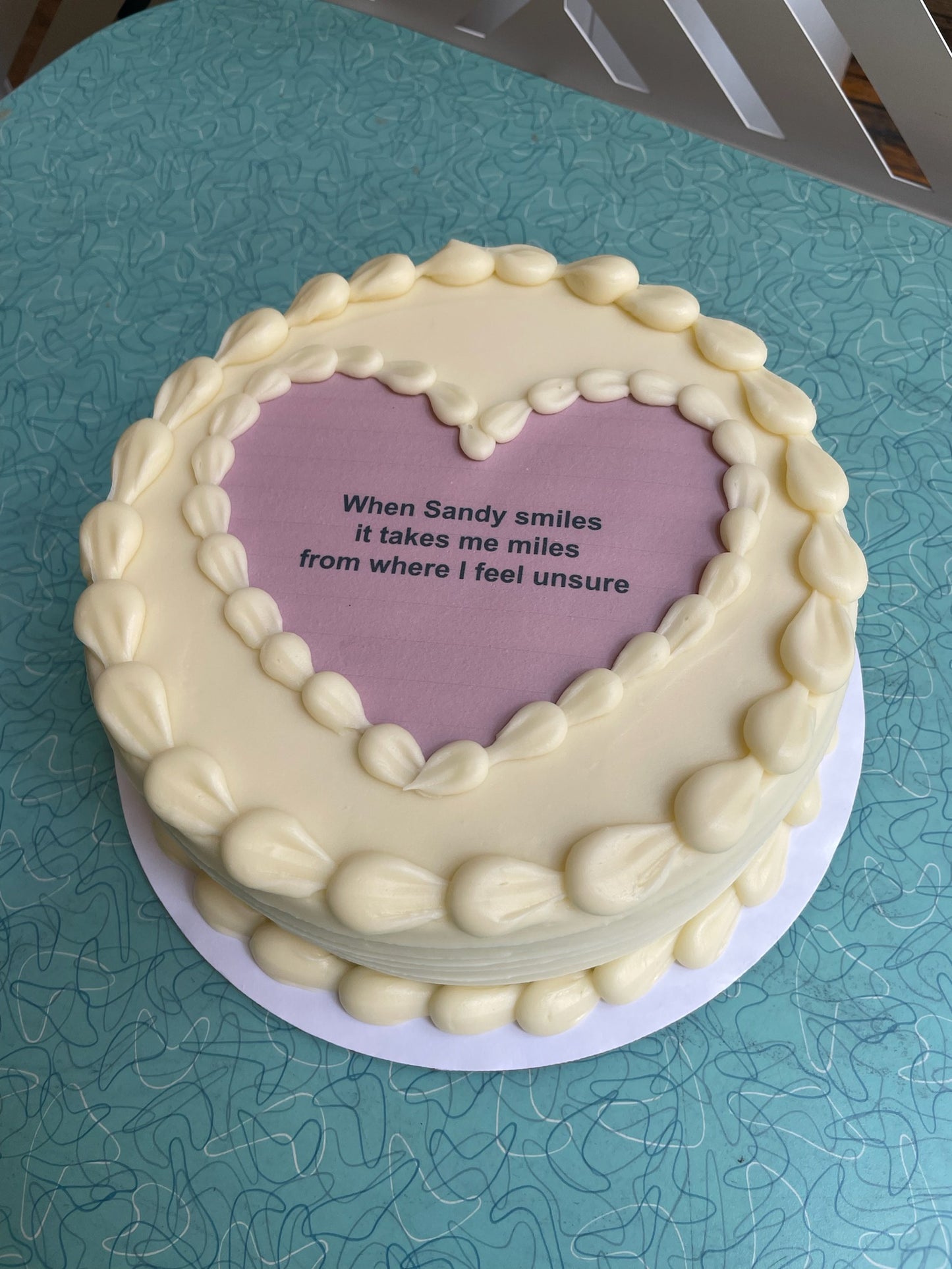 A round, pale yellow frosted cake with a large, heart-shaped purple edible image on top that reads, "When Sandy smiles it takes me miles from where I feel unsure."