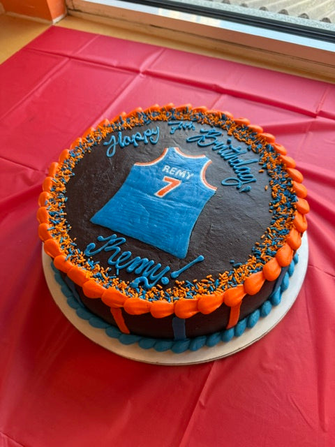 A round birthday cake decorated with dark chocolate frosting, orange and blue piped borders, and a blue basketball jersey graphic with the name 'REMY' and number 7. Blue script text on the cake reads 'Happy 4th Birthday Remy!'.