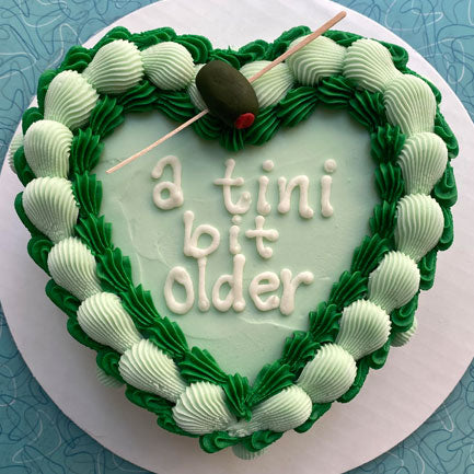 A decorated cake with green buttercream and a fondant olive topper with the text 'a tini bit older' written on it, placed on a blue surface.