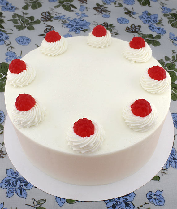 A round lemon raspberry cake with white frosting and red raspberry candy garnishes on top, placed on a plate with a floral pattern in the background.