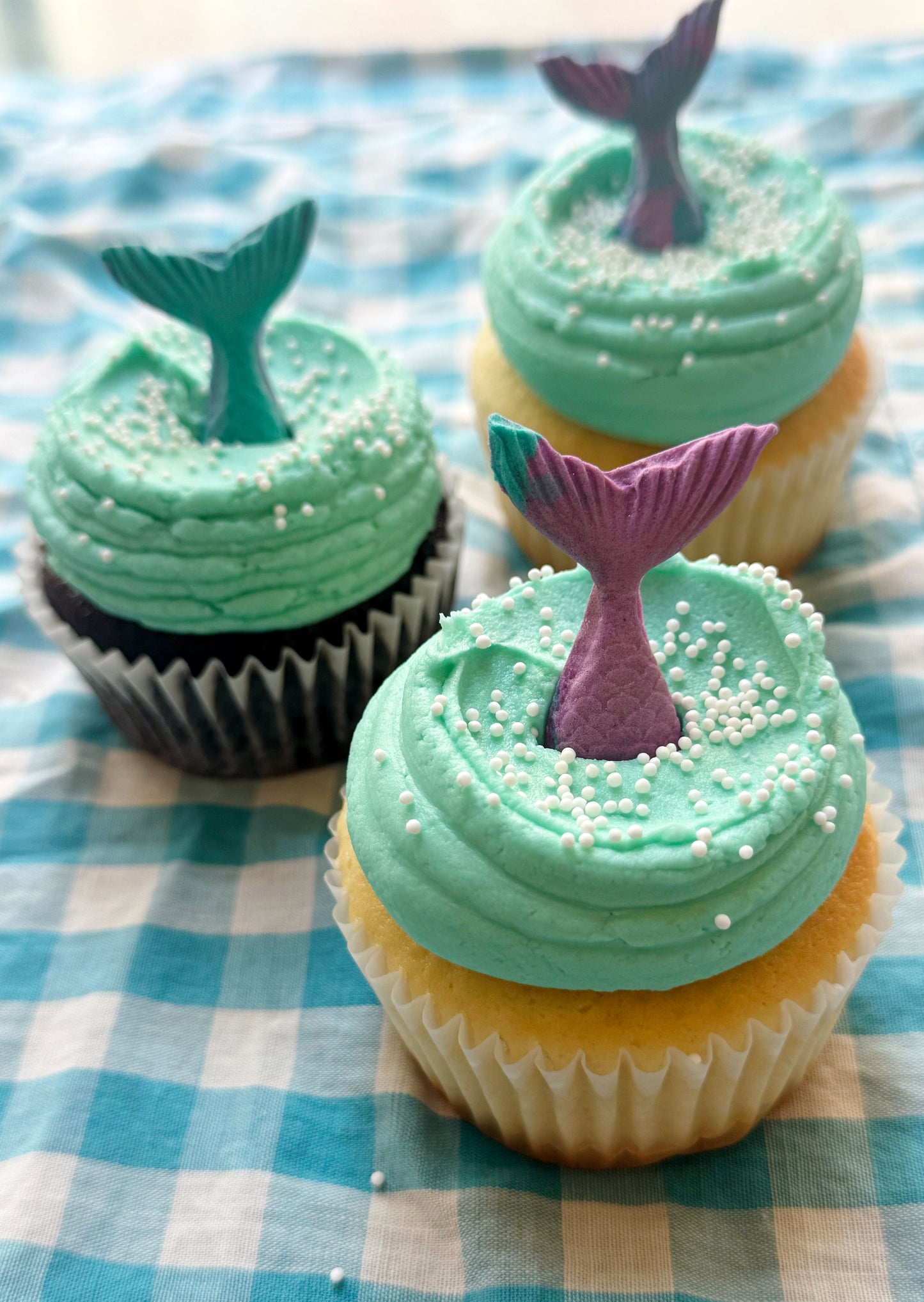 Cupcakes with green frosting and mermaid tail decorations on a checkered tablecloth.