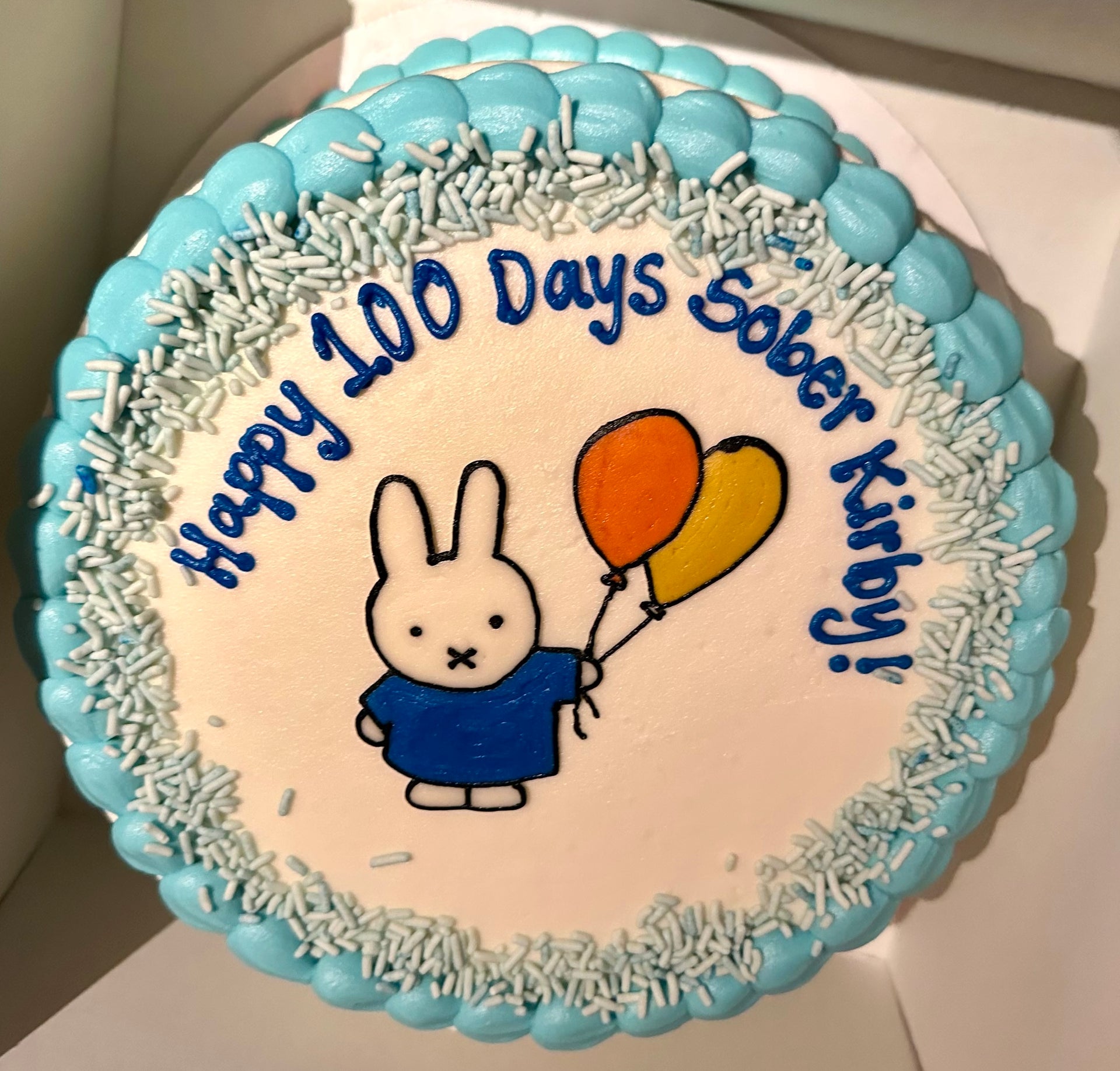 Cake with a cartoon character holding balloons and text 'Happy 100 Days Sober Kirby' on a white background.