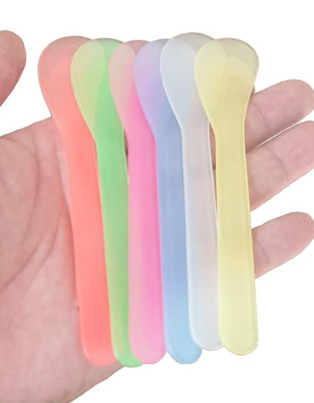 A hand holds a fan of six colorful, disposable cosmetic spatulas, including shades of orange, green, pink, purple, light blue, and yellow.