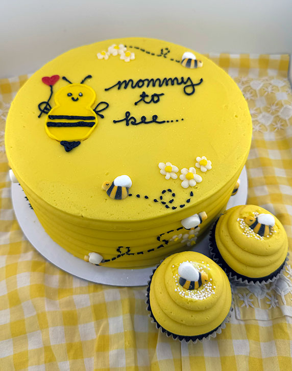 A yellow, round cake with vanilla buttercream frosting, decorated with cute edible bees and the phrase 'mommy to bee'. Accompanied by two cupcakes with the same design.