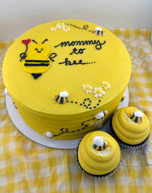 A yellow, round cake with vanilla buttercream frosting, decorated with cute edible bees and the phrase 'mommy to bee'. Accompanied by two cupcakes with the same design.