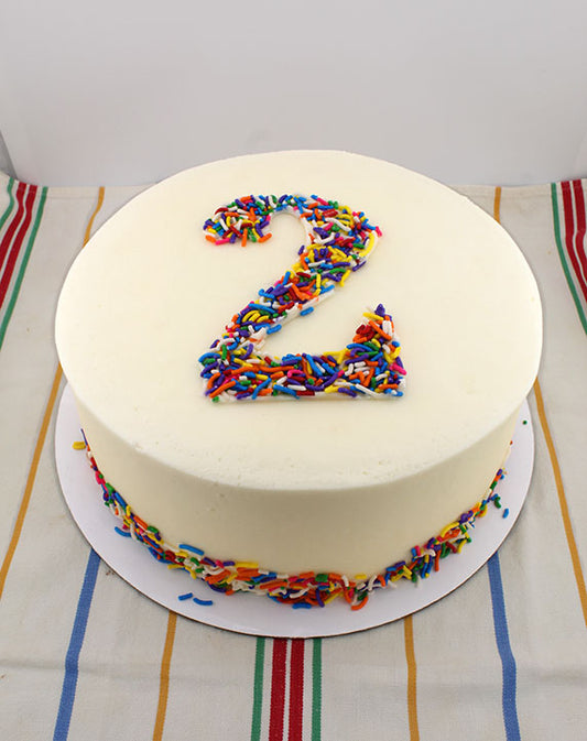 A round cake with creamy white buttercream frosting and a number '2' made of multicolored sprinkles on top, placed on a striped tablecloth.