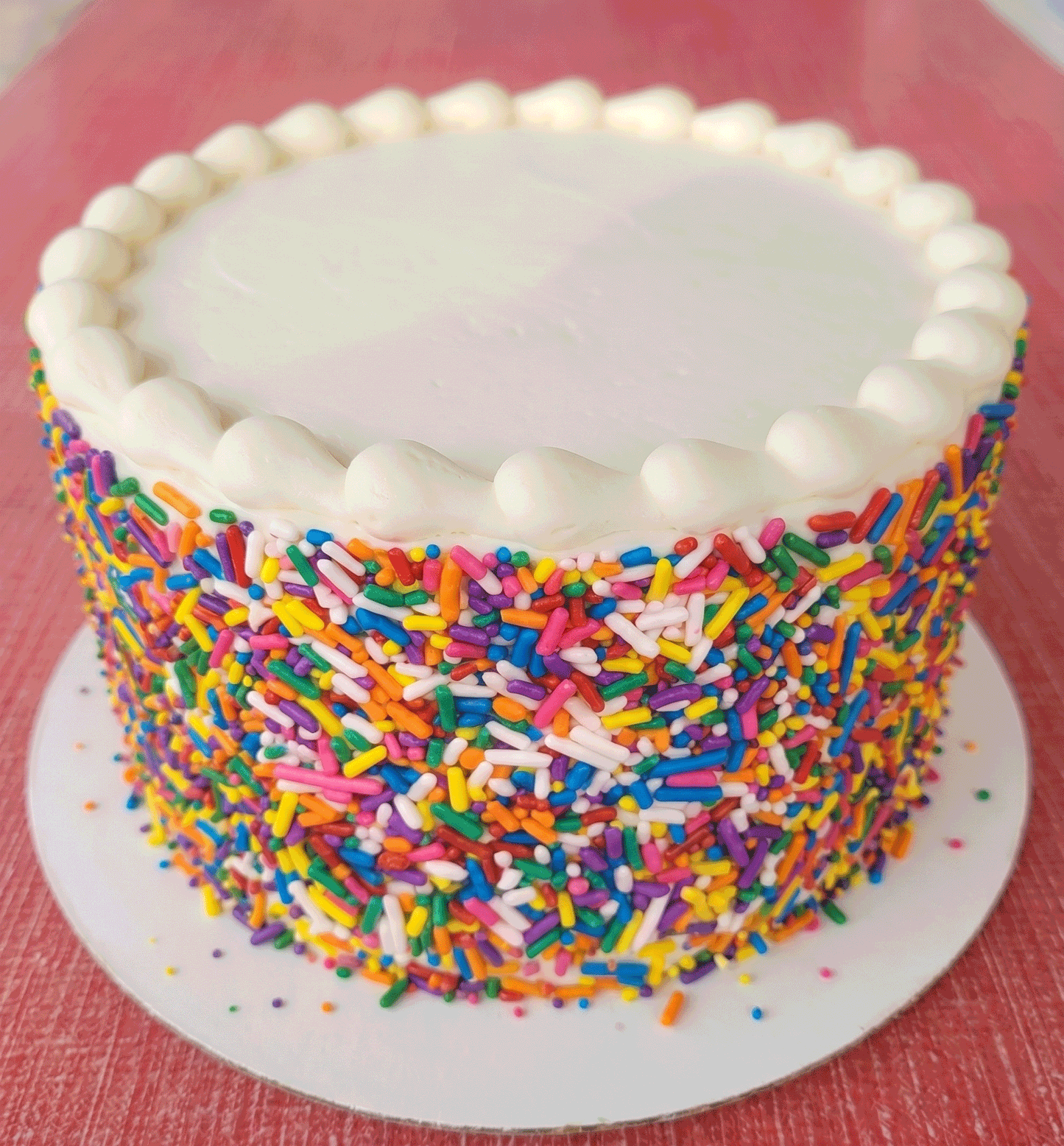 Buttercream cake with white frosting and decorative bold rainbow sprinkles on a red surface