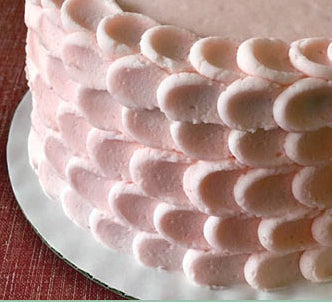 A round layer cake is decorated with soft pink buttercream in a petal or scalloped pattern, creating a textured, elegant finish. The frosting technique involves overlapping oval-shaped swipes of icing around the sides of the cake, giving it a delicate, almost floral appearance. The cake sits on a white cake board atop a deep red textured surface. Ideal for weddings, baby showers, or birthdays, this pastel pink petal cake offers a romantic and feminine aesthetic perfect for special celebrations.