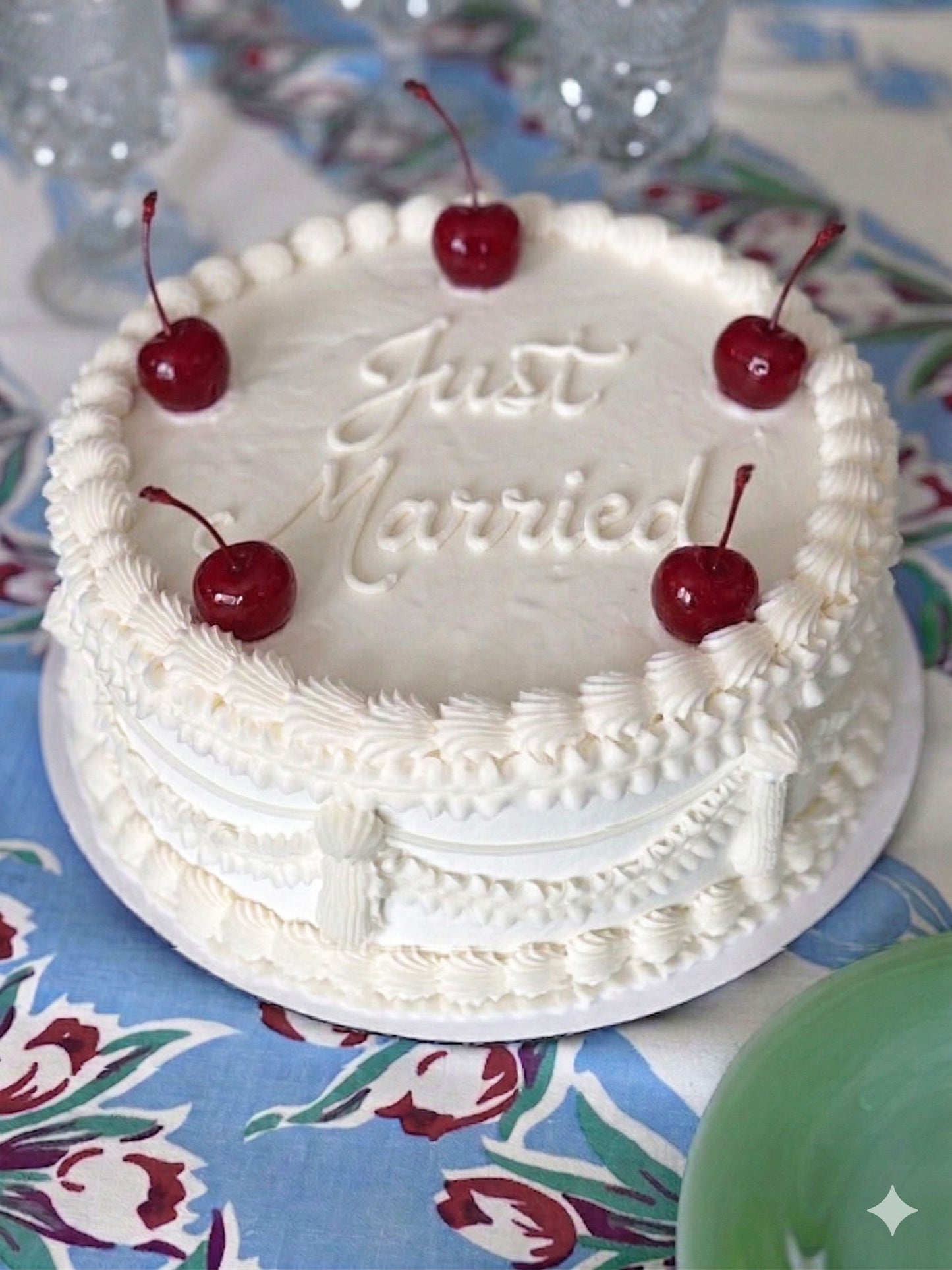White Rococo-inspired cake with 'Just Married' inscription and cherries on a floral tablecloth.