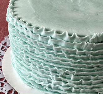 A close-up image of a cake with a ruffled, piped frosting in a light green color, possibly vanilla or lemon flavor, on a white plate.