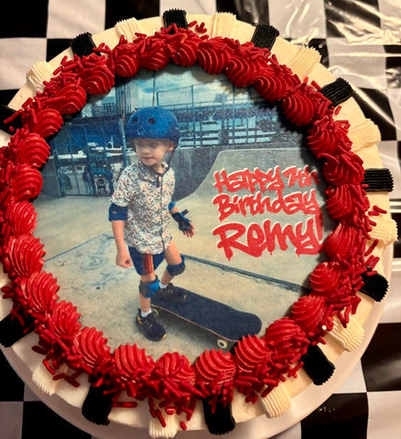 A round birthday cake decorated with red frosting swirls and black-and-white checkered edges, featuring a custom photo of a boy on a skateboard with the text 'Happy Birthday Romy!'.