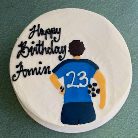 Round birthday cake with a person wearing a blue jersey with the number 23, on a green background.