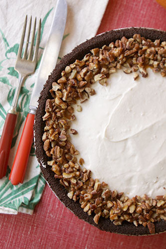 A piece of turtle cheesecake pie with a layer of caramel and crushed chocolate wafers on top, garnished with pecans and a swirl of hot fudge sauce, served on a plate with a fork.