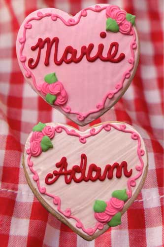 Two heart-shaped sugar cookies with personalized text. One cookie is decorated with the name 'Marie' and the other with the name 'Adam', both with pink and red decorations on a red and white checkered background.