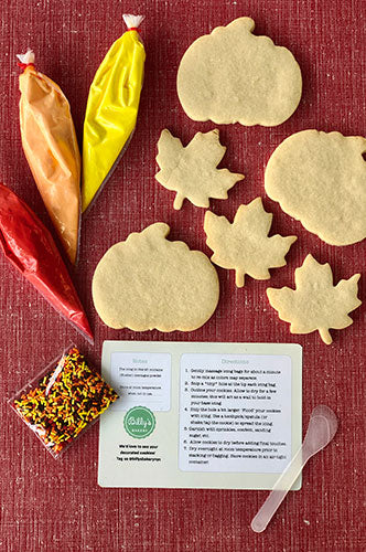 Fall-themed cookie decorating kit with pumpkin and maple leaf sugar cookies, red, orange, and yellow icing bags, autumn-colored sprinkles, and decorating instructions on a red background