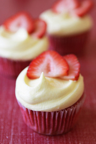 A photo of strawberry lemonade cupcakes with a lemon buttercream frosting and garnished with a strawberry slice on top, sitting on a red surface.