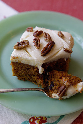 A slice of pumpkin bar cake with cream cheese icing and chopped pecans on top, served on a plate with a spoon resting beside it.