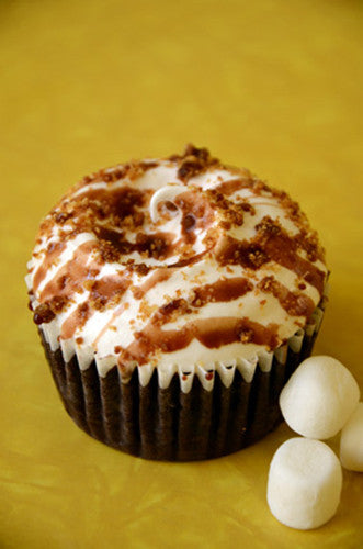 A chocolate cupcake with meringue frosting, graham cracker crumbles, and dark chocolate drizzle, presented on a yellow surface with marshmallows beside it.