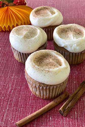 Cinnamon batter cupcakes with cream cheese frosting, dusted with cinnamon sugar, displayed on a red surface with cinnamon sticks beside them.